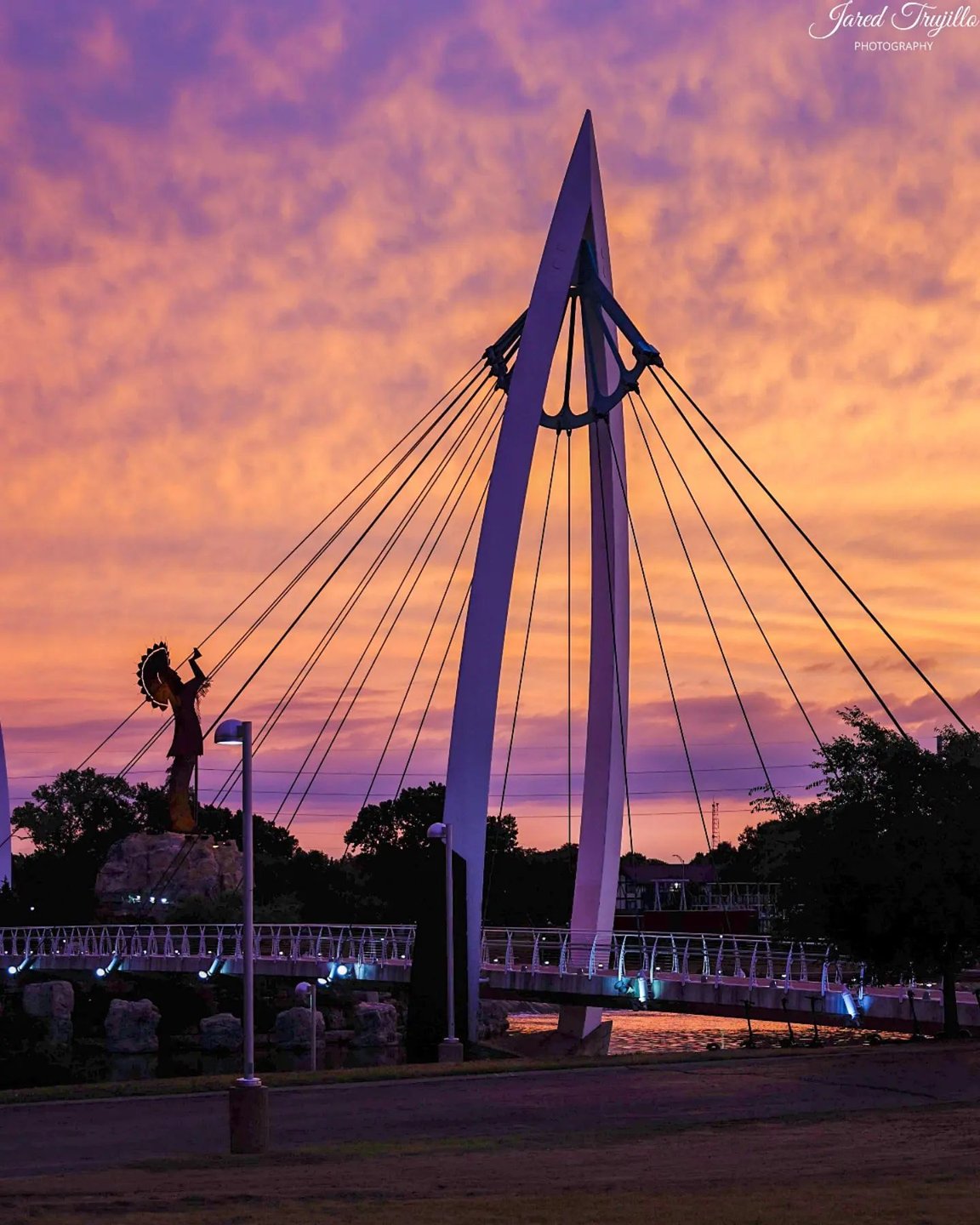 Pedestrian bridge over river at sunset with the Keeper of the Plains sculpture in the background